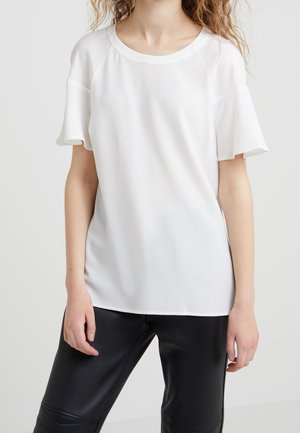 Persoon die een losse witte top met korte mouwen en ruches draagt en een zwarte leren broek, staand voor een effen achtergrond.