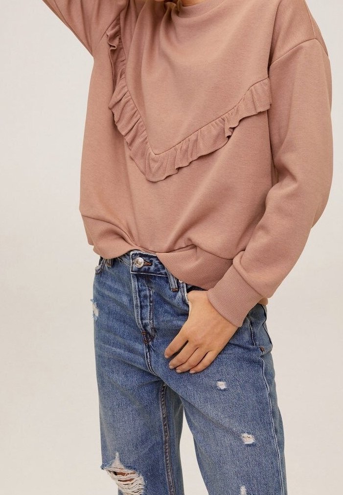 Różowy sweatshirt z falbaniastymi detalami przy dekolcie, w połączeniu z poszarpanymi niebieskimi dżinsami z wysokim stanem i z przetarciami.