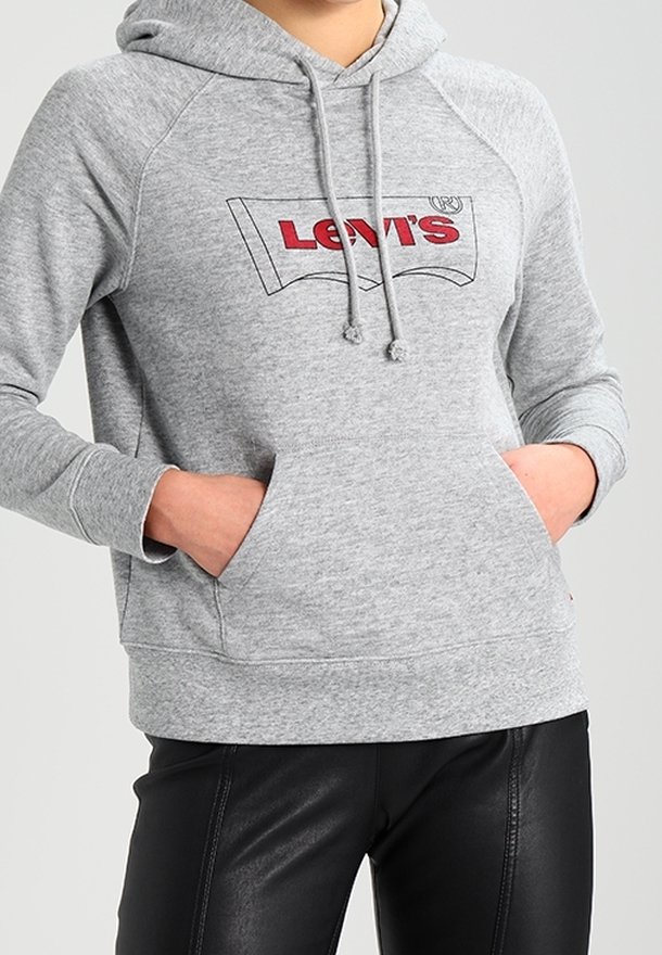 Personne portant un sweat à capuche gris Levi’s avec un logo rouge sur la poitrine, les mains dans la poche avant, associé à un pantalon en cuir noir.