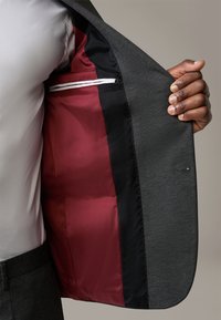 Ciemnoszara marynarka zaprezentowana rozpięta, odsłaniająca bogate bordowe satynowe podszewki z czarnym wykończeniem i białym detalem na kieszeni.