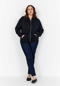 Sudadera negra con cremallera y cordones, bolsillos frontales y puños acanalados, combinada con jeans skinny azul oscuro y zapatos planos negros.
