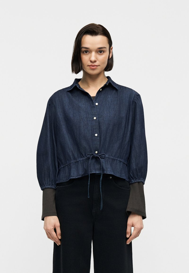 Νέα γυναίκα με κοντά σκούρα μαλλιά, φορώντας σκούρο πουκάμισο από τζιν με κουμπιά και δεμένη μέση, και μαύρο παντελόνι, στέκεται μπροστά σε λευκό φόντο.