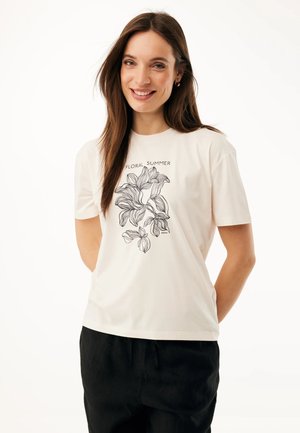 Uśmiechnięta kobieta z długimi brązowymi włosami w białej koszulce z czarnym kwiatowym wzorem i napisem "Floral Summer", ubrana w czarne spodnie.