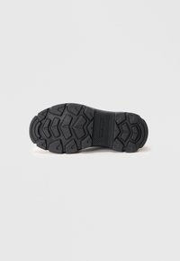 Black rubber shoe sole features deep traction patterns, a textured central grip, and a raised branding element. Flat, durable design enhances grip.