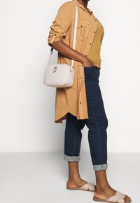 Světle hnědá košile s kabátem, hořčicový top, tmavě modré vyhrnuté džíny, růžové sandály s křížovými popruhy, malá bílá kabelka přes rameno s logem.