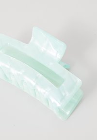 Fermaglio per capelli in acrilico verde menta con un design ondulato, dotato di un centro aperto e grandi denti per una presa sicura. Finitura liscia e lucida.