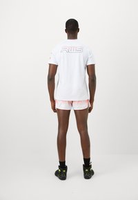 Man wearing white short-sleeve shirt with "RWS" text on back, white and red shorts, black socks, and black sneakers with neon details, standing.