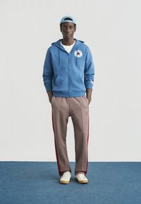 Sweat à capuche zippé bleu avec écusson logo, associé à un pantalon à carreaux marron avec des bandes latérales rouges et des baskets blanches. Look décontracté et sportif.