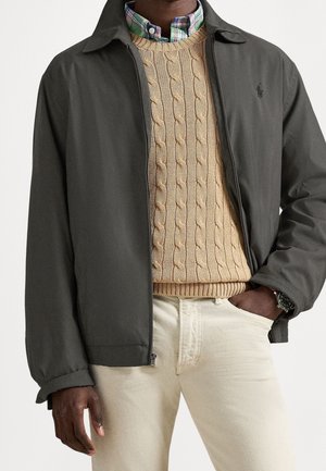 Homme portant une veste zippée gris foncé par-dessus un pull beige torsadé et une chemise à carreaux, avec un pantalon clair et une montre-bracelet.