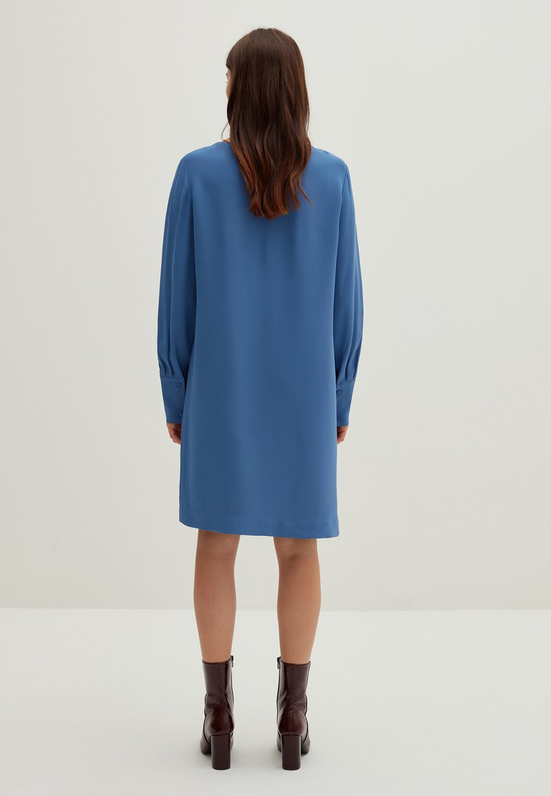 Abito lungo fino al ginocchio con maniche lunghe in tessuto blu uniforme. Presenta una vestibilità comoda, polsini con bottoni e una texture liscia e leggera. Indossato con stivaletti marroni alla caviglia.