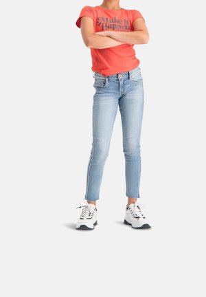 Camiseta roja con el texto "Haz que suceda", jeans ajustados azul claro con rayas laterales y zapatillas blancas con detalles en negro.