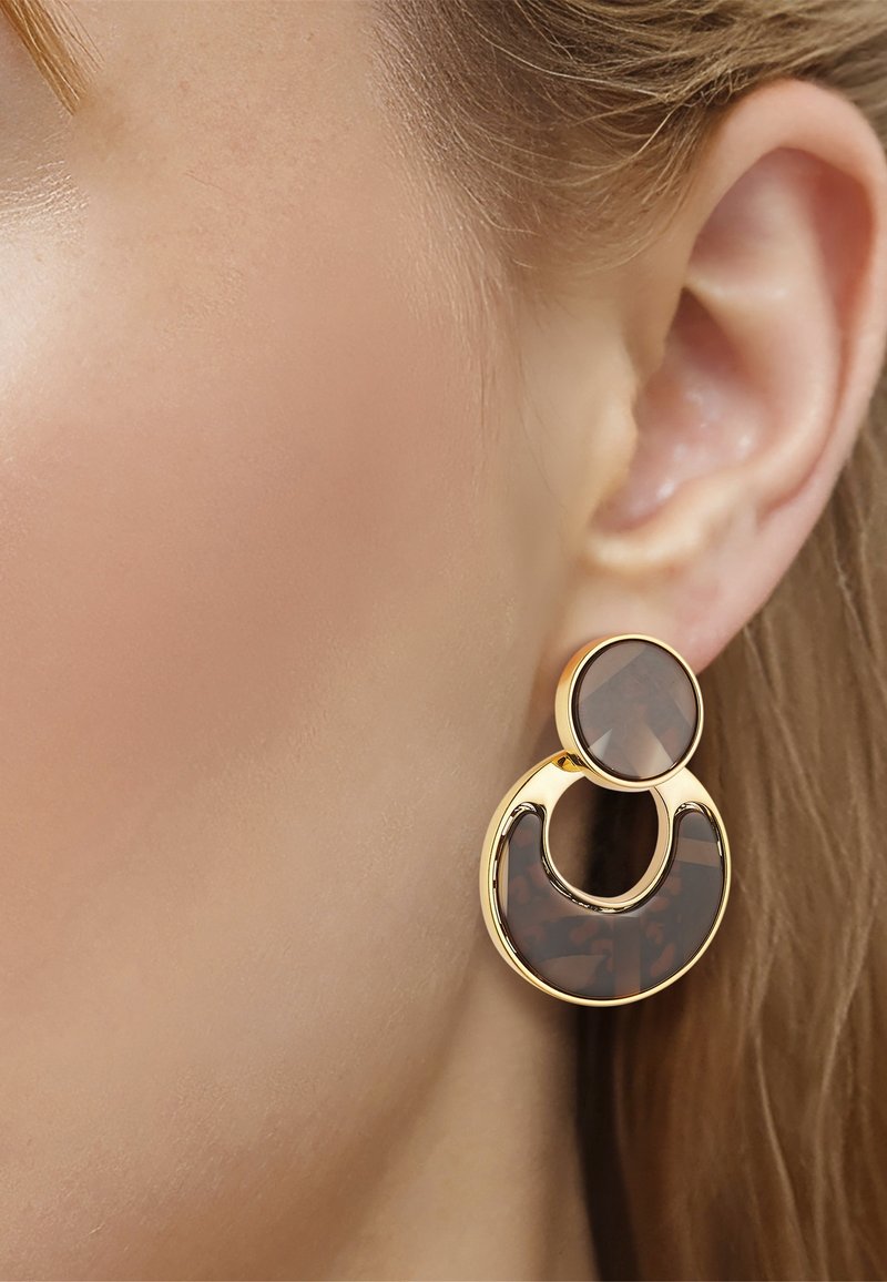 Nahaufnahme eines Frauenohres, das einen goldenen, schildpattgemusterten, runden Tropfen-Ohrring trägt.