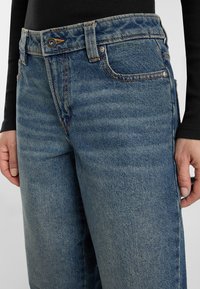 Close-up of a person wearing faded blue denim jeans with orange stitching and a black long-sleeve top, hands resting by sides.