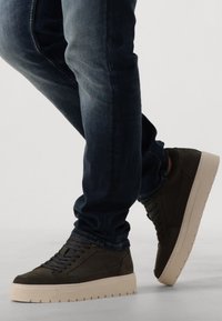 Donkergroene sneakers met textuurpanelen, beige rubberen zool en ronde veters, gecombineerd met donkere spijkerbroeken.