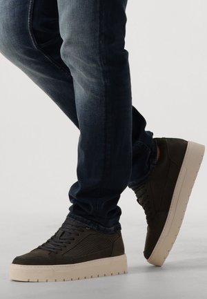 Baskets vert foncé avec des panneaux texturés, semelle en caoutchouc beige et lacets ronds, assorties à un jean en denim foncé.