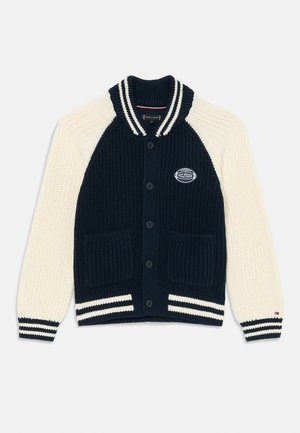 Gilet tricoté avec un corps bleu marine et des manches crème, présentant des poignets côtelés, deux poches avant et un écusson logo sur la poitrine.
