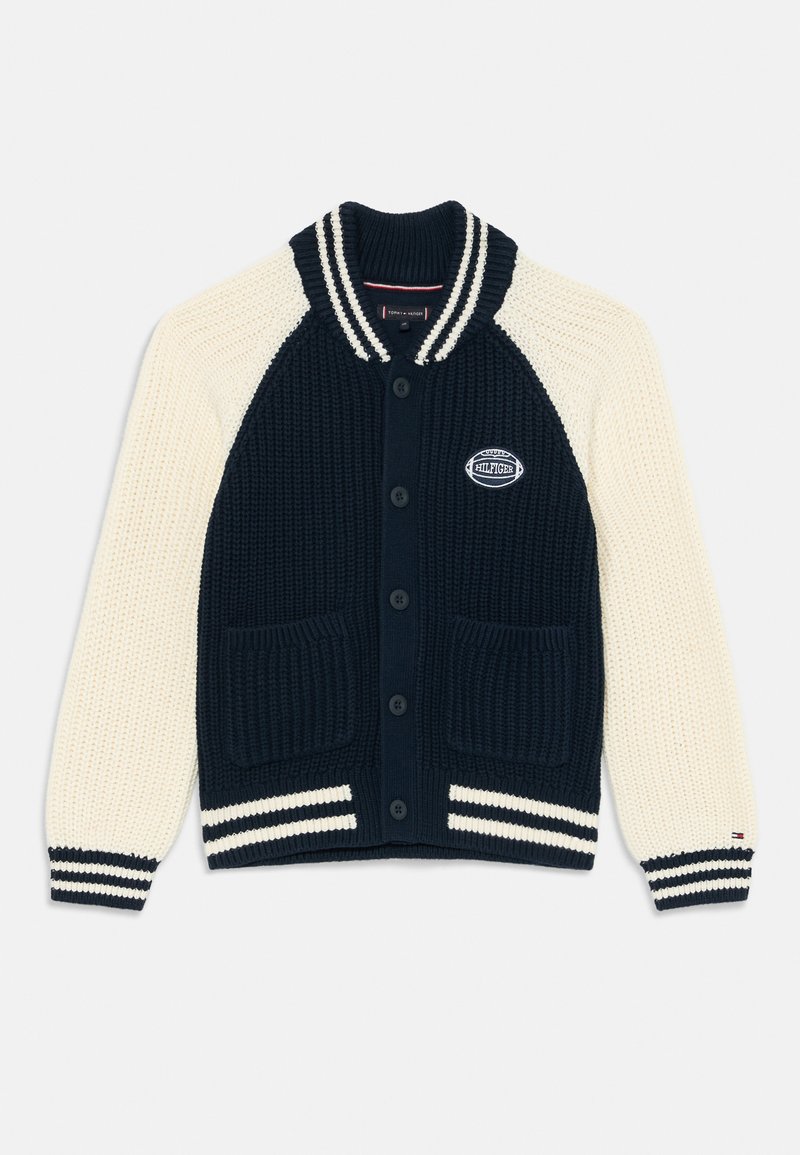 Cardigã de malha com corpo azul-marinho e mangas creme, apresentando punhos canelados, dois bolsos frontais e um emblema no peito.