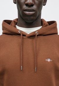 Barna kapucnis pulóver pamut keverékből, állítható kapucnival, ezüstszínű fémalkatrészekkel és hímzett logóval a mellrészen. Sima tapintású, kényelmes, bő fazon.