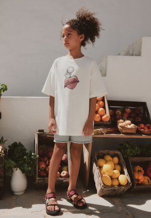 Barn med krøllet hår iført en hvid t-shirt og denimshorts står nær kurve og kasser med frugt og grøntsager udendørs.