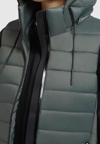 Smanicato imbottito di colore verde scuro, con collo alto, cerniera nera lucida e cordino regolabile. Tessuto morbido con motivo trapuntato.