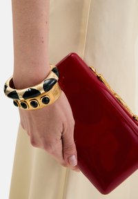 Kultainen ja musta rannekoru, jossa on tekstuurillisia yksityiskohtia, sijoitettuna kädelle, joka pitää kiiltävää punaista kirjekuorilaukkua kultaisella ketjulla.