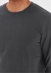 Hombre vestido con una camiseta de manga larga gris oscuro, que muestra la parte superior del pecho y los hombros, contra un fondo liso.
