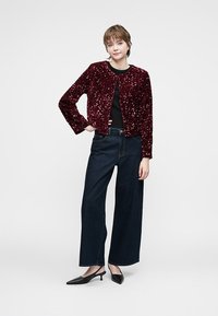 Jauna sieviete stāv ar roku uz gurniem, ģērbusies bordo krāsas sequīnu jaku, melnu blūzi, platām tumšām džinsiem un melniem šaurajiem zābakiem.