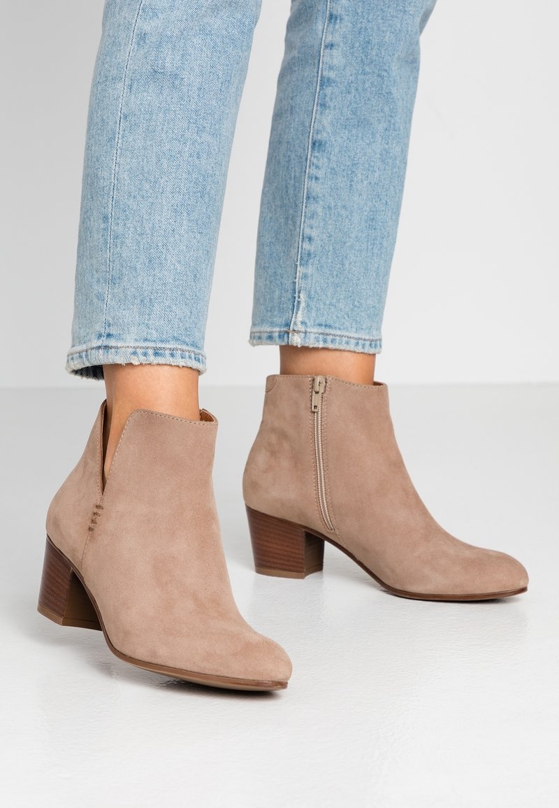 Bottes basses en daim marron clair avec fermetures éclair latérales et talons carrés portées avec un jean court bleu clair sur un sol blanc lisse.