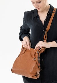 Borsa a tracolla in pelle color tan con una texture morbida, tracolla regolabile e dettagli decorativi. Apertura superiore con chiusura a cordoncino e accenti metallici.