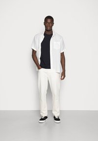 Weißes Kurzarmhemd über einem schwarzen T-Shirt, kombiniert mit weißen Hosen und schwarzen Sneakern. Das Model steht frontal und hat die Hände in den Taschen.
