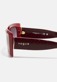 Occhiali da sole bordeaux con montatura spessa e quadrata. Le aste texturizzate presentano intricati motivi di rose e il marchio dorato "vogue". Lenti sfumate.