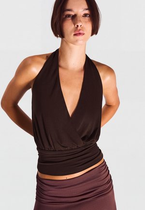 Frau mit kurzen dunklen Haaren, die ein ärmelloses schwarzes Neckholdertop und einen dunkelvioletten Rock vor einem schlichten hellen Hintergrund trägt.