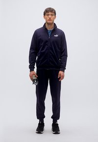 Σκούρο μπλε φόρμα από απαλό ύφασμα, που αποτελείται από τζάκετ με φερμουάρ και παντελόνι που ταιριάζει, συνδυασμένη με μαύρα αθλητικά παπούτσια και παγούρι νερού.