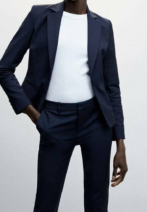 Costume sur mesure bleu marine à coupe slim, comprenant un blazer à un bouton et un pantalon à plis plats. Porté avec un haut blanc côtelé.