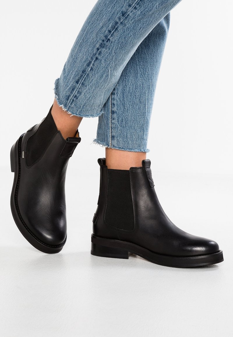 Bottines en cuir noir portées avec un jean court en denim bleu clair sur fond blanc.