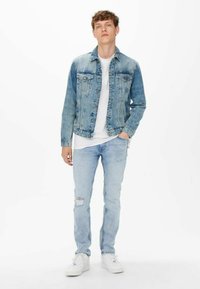 Light blue denim jacket with front pockets and faded design, worn over a white T-shirt. Paired with light blue distressed jeans and white sneakers.