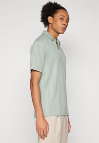 Camisa polo em tecido verde-claro com um padrão de diamantes texturizado, mangas curtas e colarinho pontiagudo. Usada com calças bege claro.
