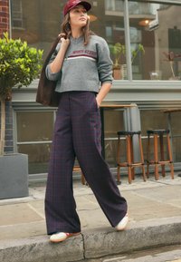 Pulover scurt gri cu textul "SPORTS STUDIO", pantaloni cu talie înaltă, largi, din material tartan în nuanțe de albastru închis și burgund, și adidași albi cu accente roșii.