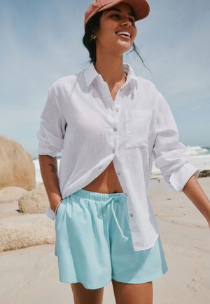 Kvinde smiler på en strand iført en hvid skjorte med knapper, lyseblå shorts og en lyserød kasket med klipper og havets bølger i baggrunden.