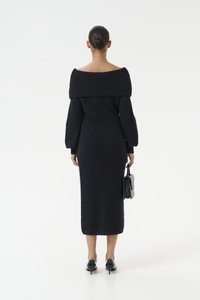Lungo vestito nero lavorato a maglia con scollo a spalle scoperte, maniche a sbuffo e silhouette aderente, abbinato a una piccola borsa nera.