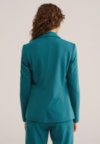 Bermudski suknjič v modrozelene barve s krojenim prileganjem, dolgimi rokavi in zarezami na ovratnici. Gladek, teksturiran material z vgrajenim skritim zadnjim razporkom.