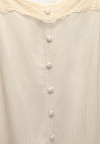 Blouse crème avec six boutons ronds recouverts de tissu le long du centre et une bordure en dentelle autour de l'encolure.