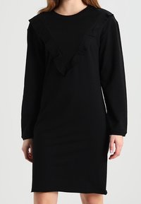 Svart långärmad klänning med V-formad halsringning prydd med volangdetaljer. Tillverkad av mjukt tyg, knälång med avslappnad passform.