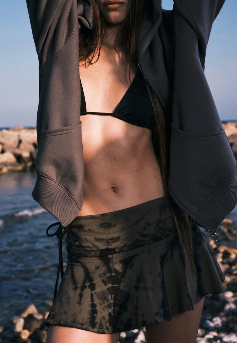 Sweat à capuche gris, haut de bikini noir et mini-jupe en tie-dye sombre. La tenue présente des textures douces et des formes fluides, avec une coupe décontractée.