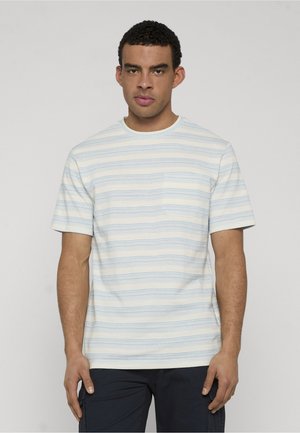 Mand iført lyseblå og hvide stribede kortærmede skjorte med brystlomme og mørke bukser, stående mod en ensfarvet baggrund.