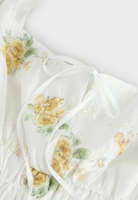 Tissu blanc transparent avec broderie florale jaune et fines attaches, froncé à la couture, probablement partie d'un vêtement ou détail de robe.