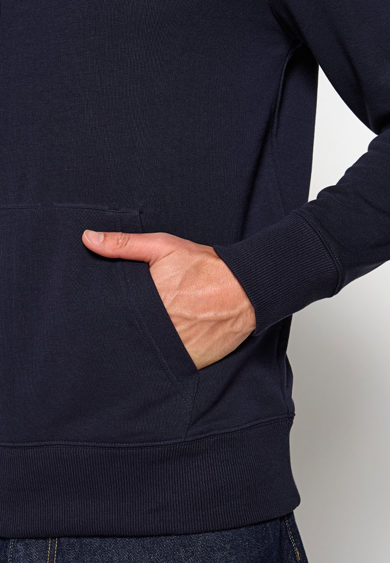 Main dans la poche frontale d'un sweat à capuche bleu marine, montrant des détails de couture sur les poignets à côtes et la poche.