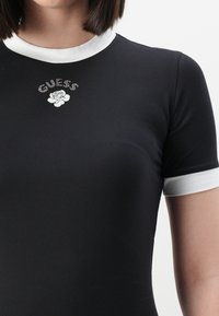 Svart tettsittende t-skjorte med hvit krage og ermtrim. Har rhinestein «GUESS»-tekst og en hvit rosegrafikk under. Glatt stofftekstur.