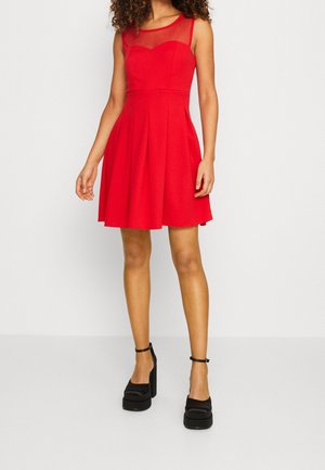 Kobieta w czerwonej sukience bez rękawów do kolan z przezroczystym panelem na górnej części dekoltu, w połączeniu z czarnymi platformowymi szpilkami na kostkę z paskiem.