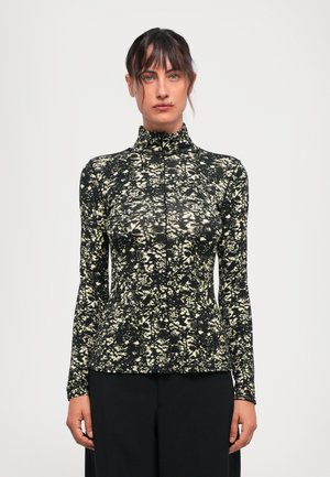 Woman with dark hair in a high-neck, long-sleeve black top with white floral pattern, standing against a plain light background.